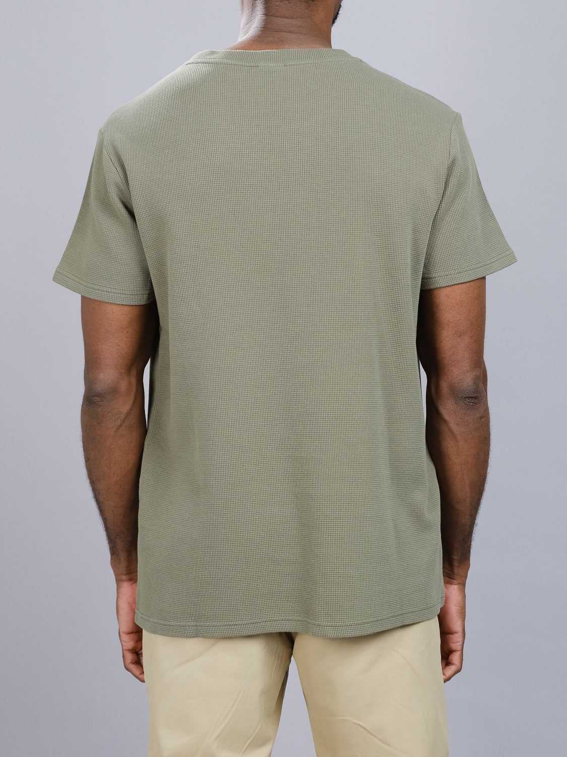 A person is seen from behind wearing an ID Menswear Urban Waffle Knit Tee in olive green and beige pants, set against a plain gray background.