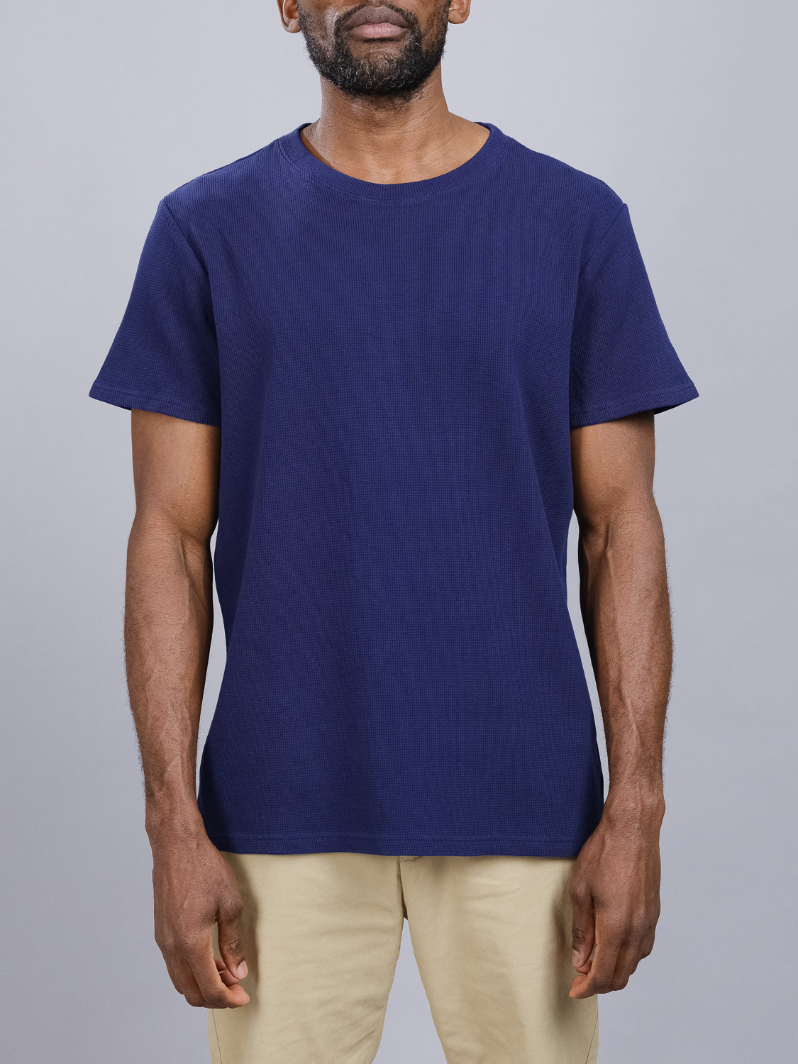 A person in an Urban Waffle Knit Tee by ID Menswear, styled in casual navy blue, pairs it with beige pants against a light gray background. The image centers on the torso with relaxed arms, capturing its simple and minimalistic style.