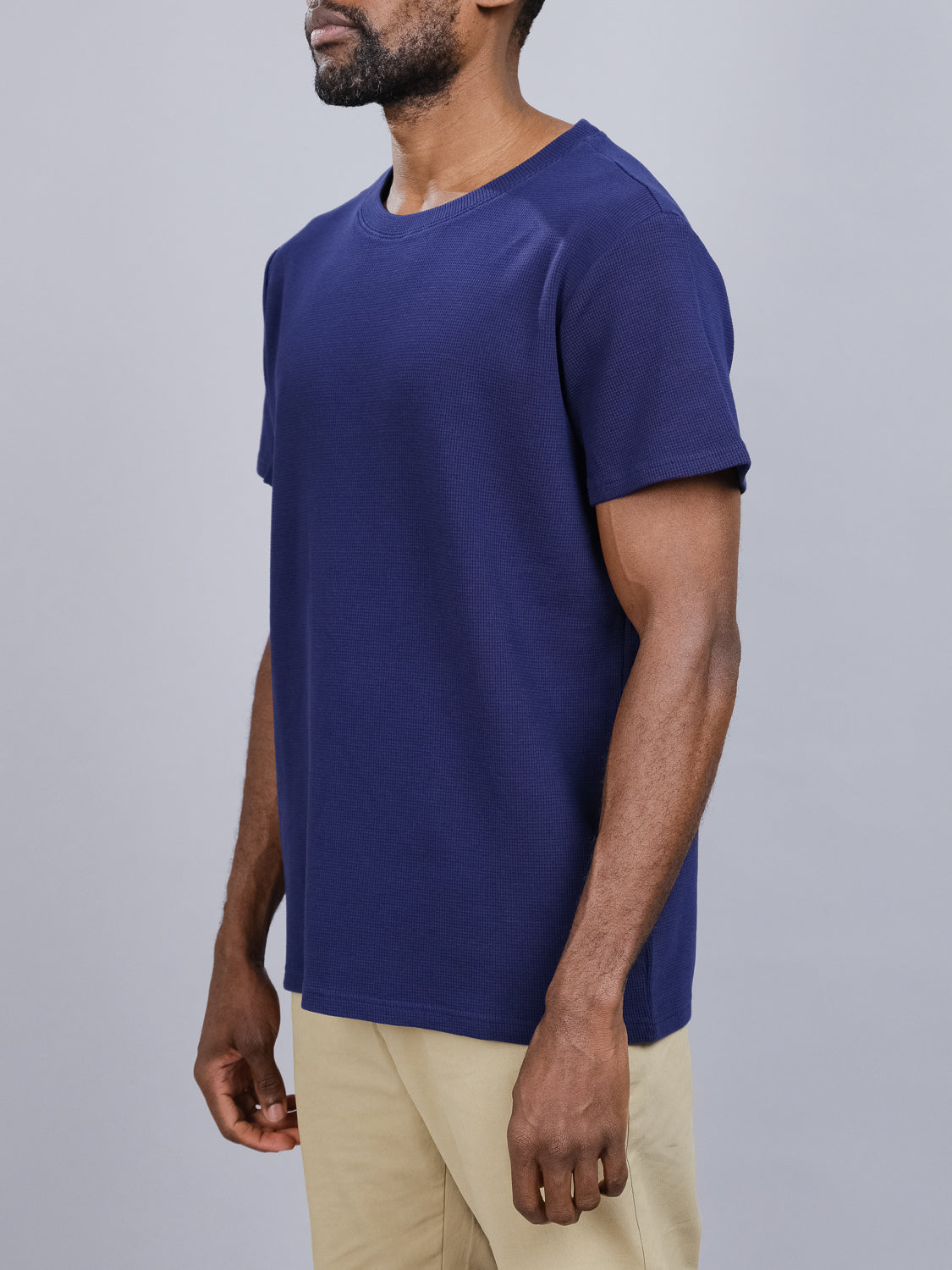 A man wears an Urban Waffle Knit Tee by ID Menswear in plain navy blue with beige pants against a gray background, focusing on the torso and arms to emphasize the casual look of the cotton-spandex blend fabric.