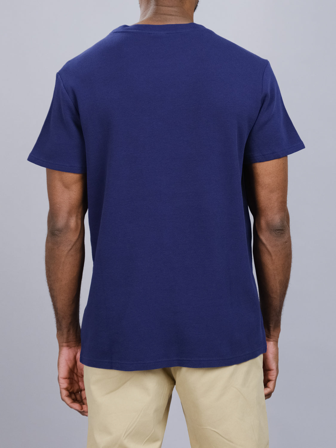 A person faces away from the camera, wearing ID Menswears Urban Waffle Knit Tee in navy blue—a comfy cotton-spandex blend—and beige pants, set against a plain gray backdrop.