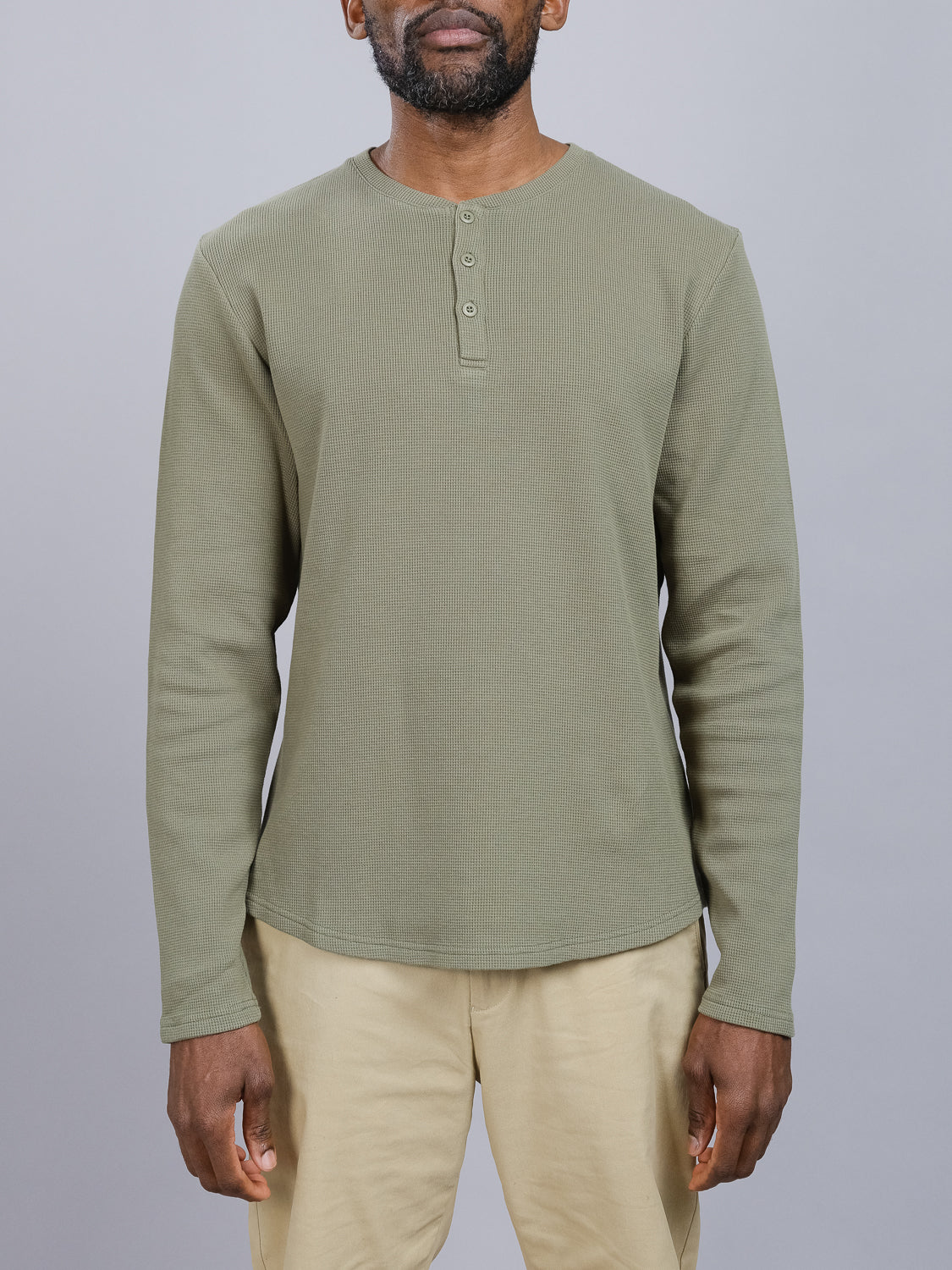 A person wears the Freestyle Waffle Knit Henley by ID Menswear, featuring a regular fit and long sleeves in olive green. Its made from a cozy cotton-spandex blend with button detailing and is paired with beige pants, set against a plain gray background.