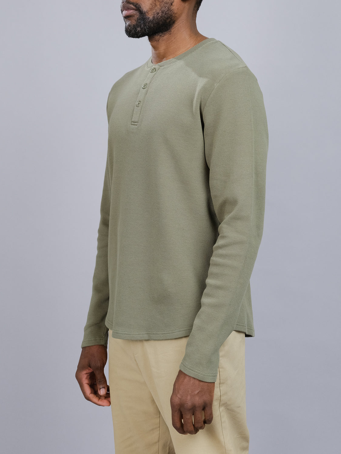 A person models ID Menswears Freestyle Waffle Knit Henley in sage green and beige pants against a gray backdrop. The regular fit shirt, made from a cotton-spandex blend, has a buttoned placket at the collar. They stand looking to the side.