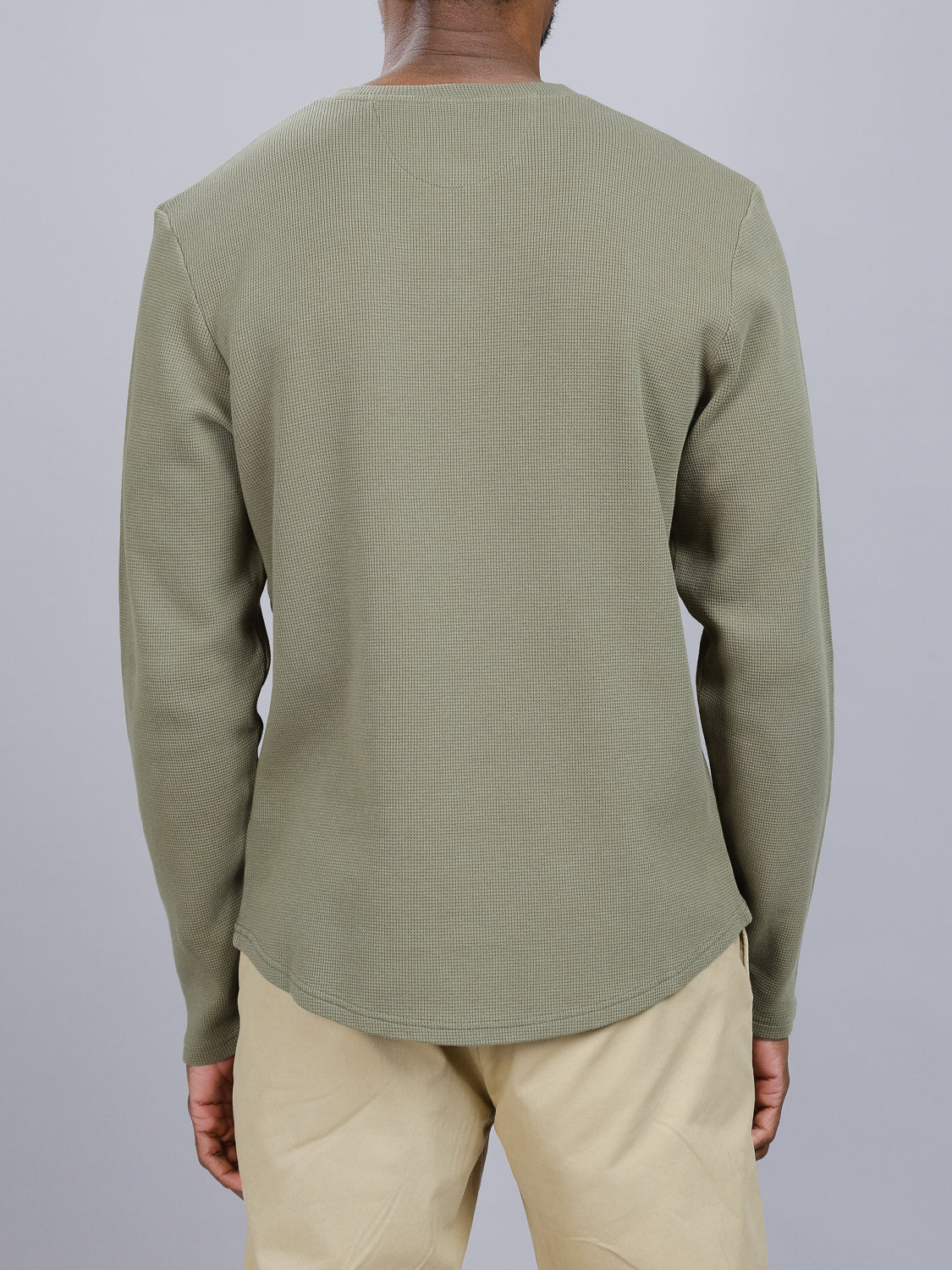 A person wearing the Freestyle Waffle Knit Henley from ID Menswear in olive green ribbed cotton-spandex and beige pants stands facing away against a gray background. The rounded hemline of the shirt provides a regular fit, enhancing the overall look effortlessly.