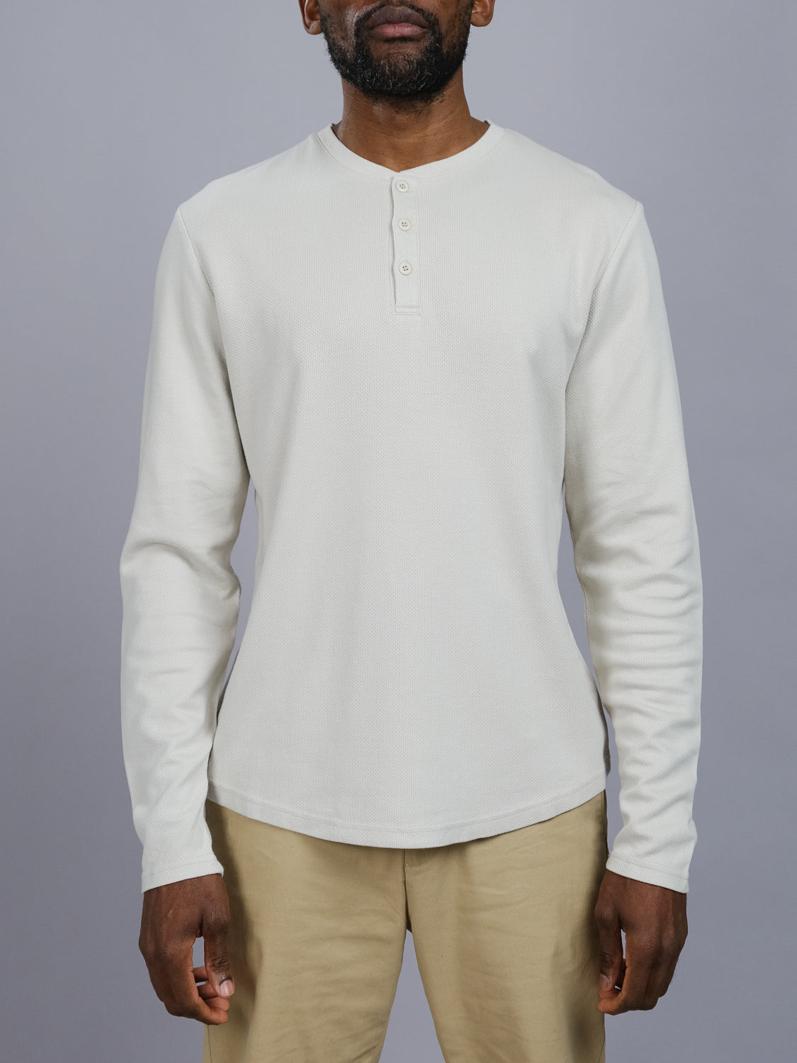 A person models the cream Freestyle Honeycomb Knitted Mesh Henley by ID Menswear and beige pants against a gray backdrop. The henley features a short buttoned placket at the neck, with a focus on knit detail from shoulders to mid-thigh.
