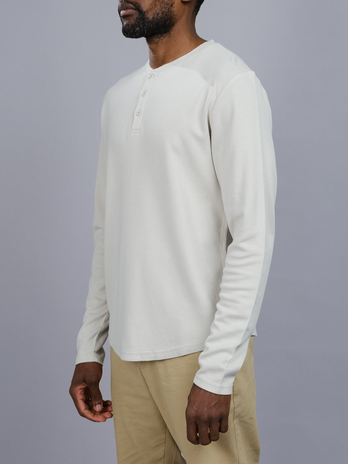 A person in a light gray Freestyle Honeycomb Knitted Mesh Henley from ID Menswear and beige pants poses against a plain gray backdrop. The shirts buttoned collar and design add casual sophistication as they turn slightly left.