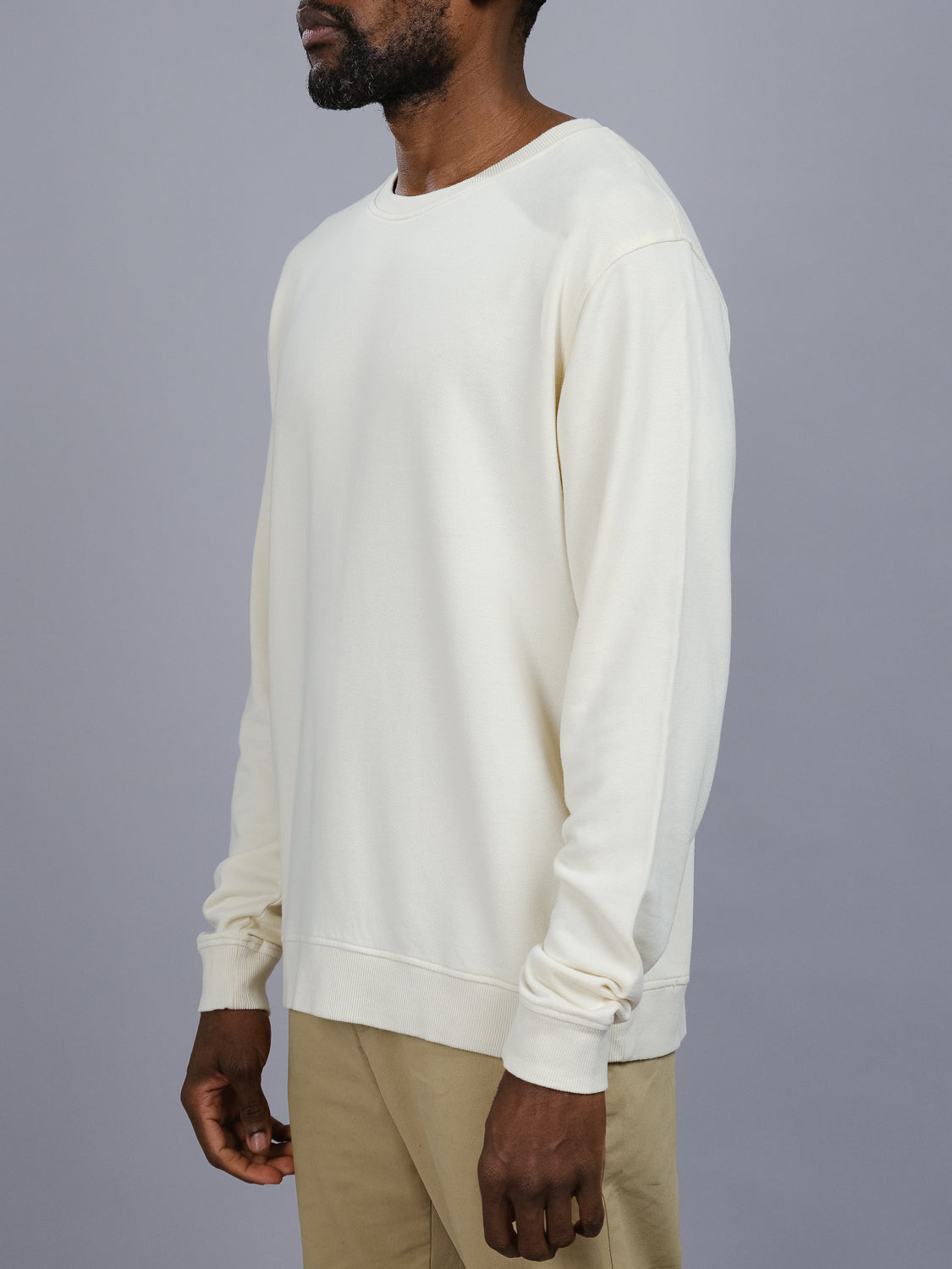 A person stands sideways in an ID Menswear Champ Bamboo Organic Cotton Sweatshirt, cream-colored, against a gray background. Only their upper body is visible, highlighting the sustainable style and natural texture of the organic cotton.