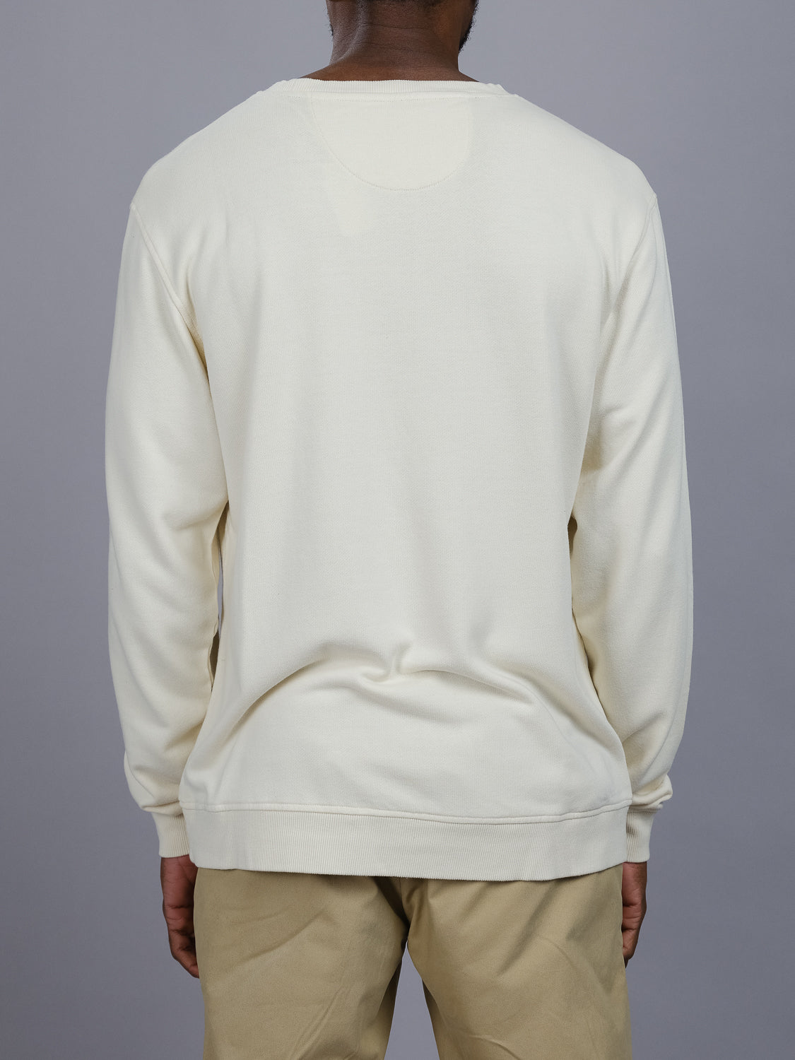 A person wearing the Champ Bamboo Organic Cotton Sweatshirt in light cream vintage wash by ID Menswear and tan pants stands with their back to the camera against a plain gray background.