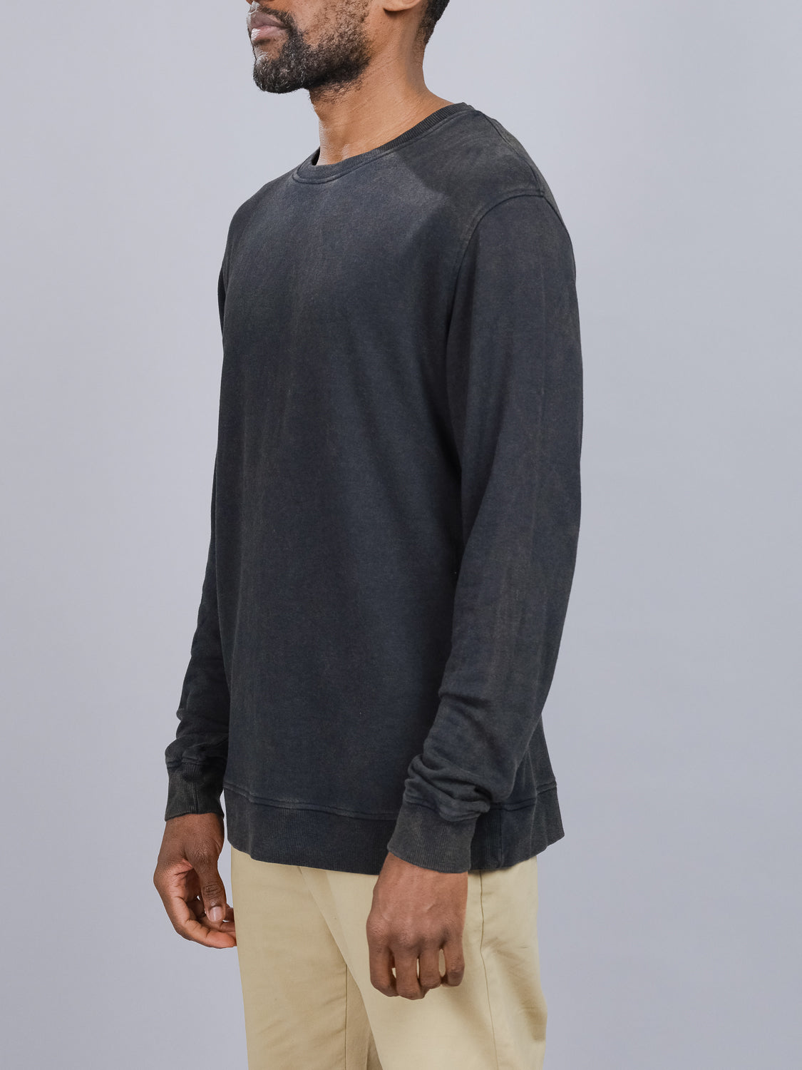 A person is wearing an ID Menswear Champ Bamboo Organic Cotton Sweatshirt in vintage wash faded black and beige pants, standing against a plain gray background. They face slightly to the left with arms relaxed at their sides.