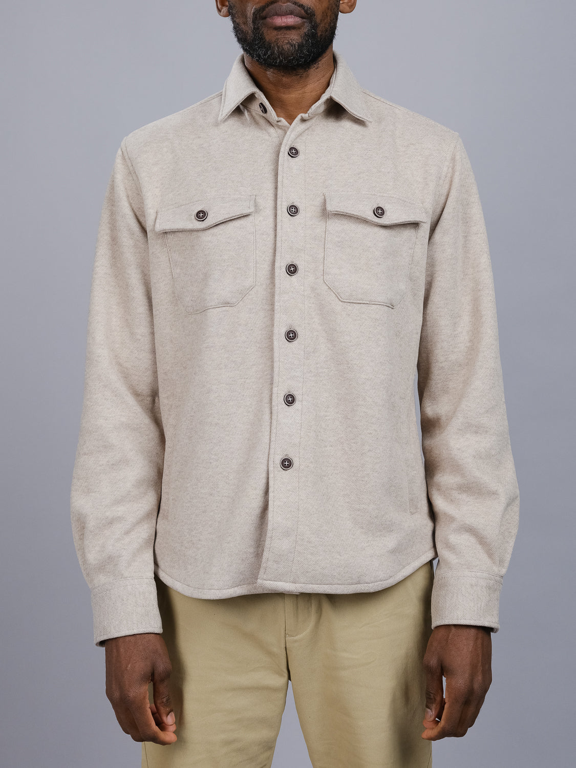 A person wearing a beige ID Menswear Yukon Shirt Jacket with two front pockets stands against a gray background. Ideal for casual outings and transitional weather, it pairs well with light khaki pants, while the dark buttons provide contrast as they stand arms relaxed by their sides.