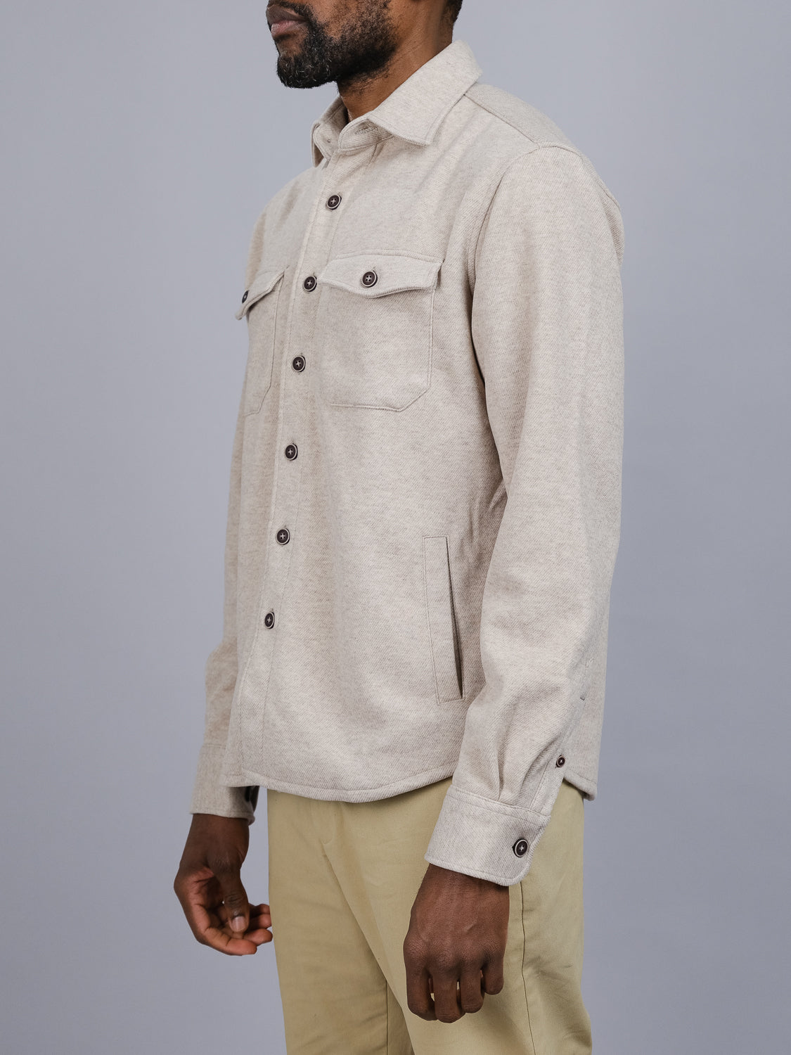 Against a plain gray background, a person models the ID Menswear Yukon Shirt Jacket in light beige with two chest pockets and dark buttons for contrast. They pair it with tan pants while facing slightly left, creating an ideal look for transitional weather and casual outings.