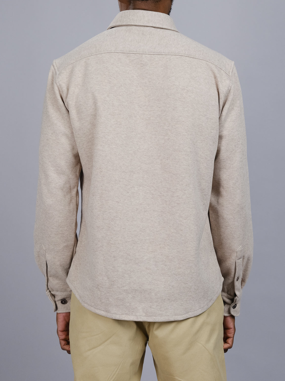 The image displays someone from behind wearing a beige ID Menswear Yukon Shirt Jacket and light pants, ideal for transitional weather, set against a plain gray background.