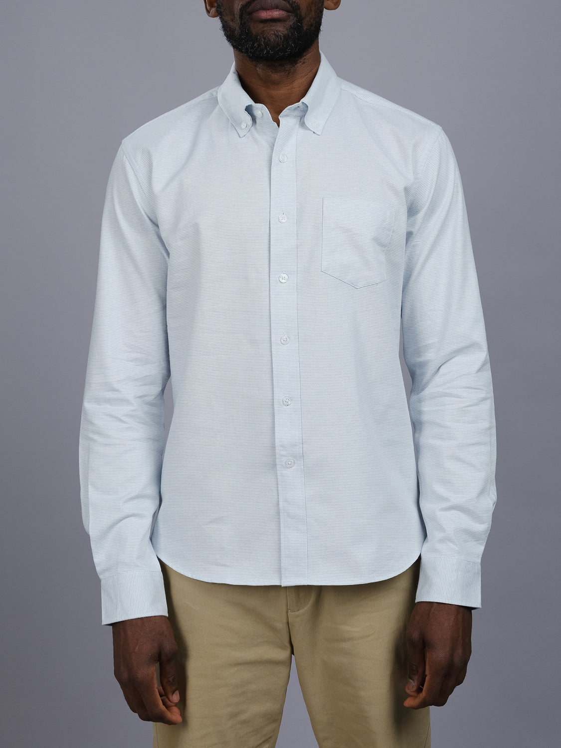 Wearing the Akira Cotton Button Down Shirt by ID Menswear in light blue, a person stands against a gray background. The shirt features a button-down collar and chest pocket and is paired with beige pants. The individuals face remains unseen.