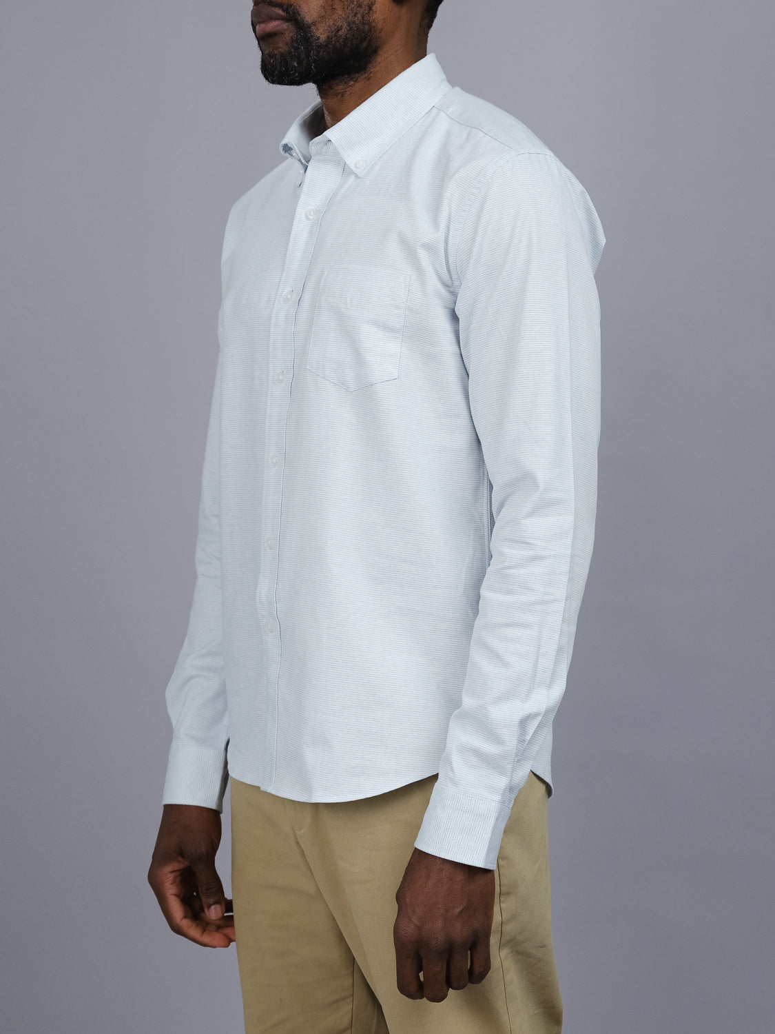 A person wearing an Akira Cotton Button Down Shirt from ID Menswear, light blue in color, with khaki pants stands against a gray background, viewed from the side with arms relaxed.