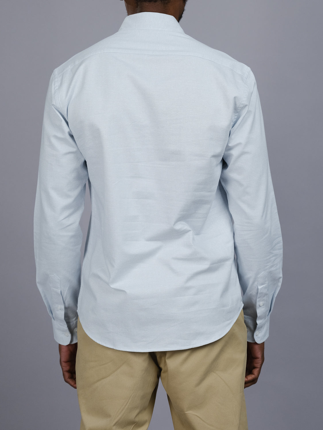 An individual in an Akira cotton button-down shirt by ID Menswear, featuring a light blue color and regular fit, pairs it with beige pants and stands facing away against a plain gray backdrop.