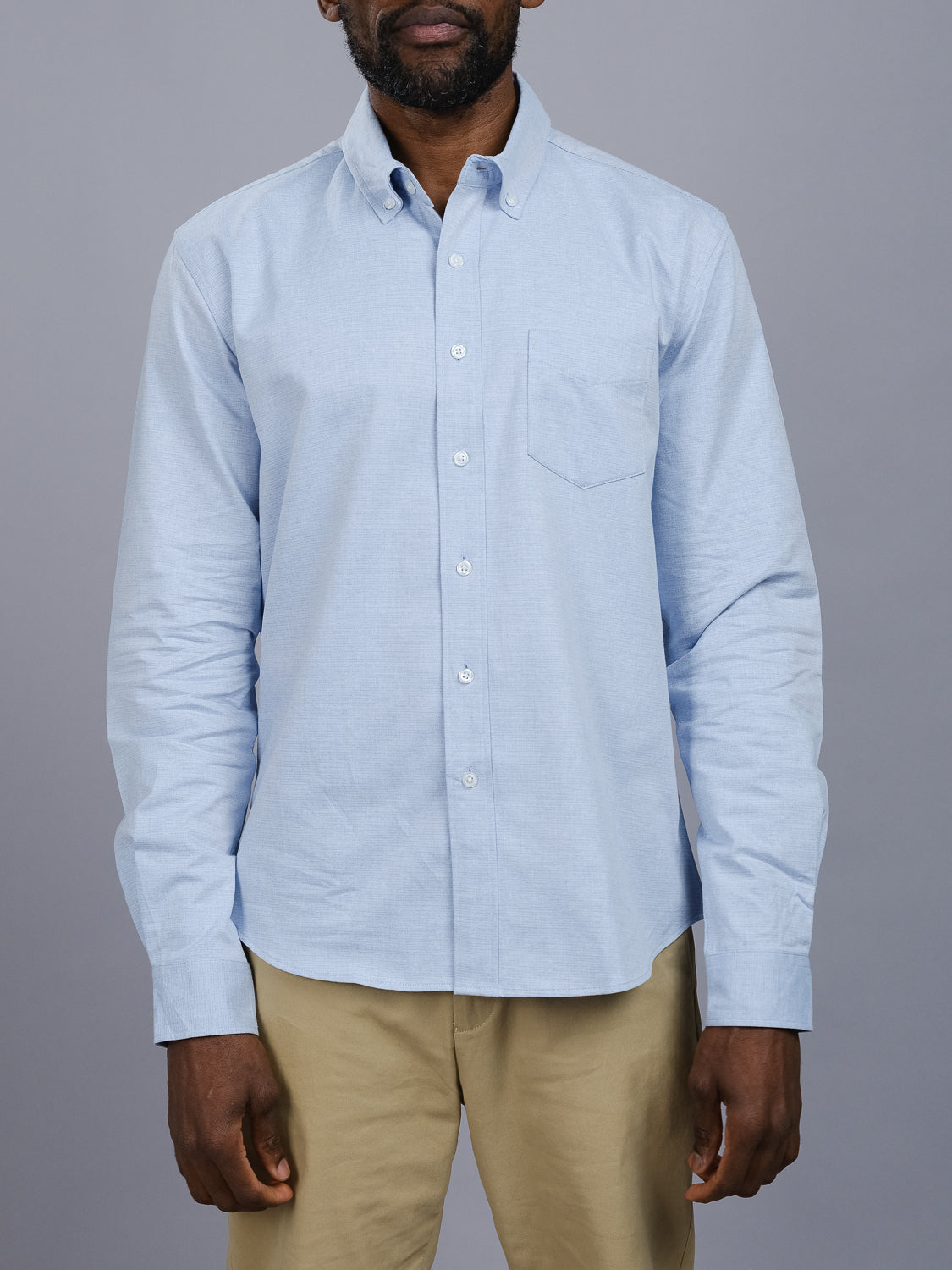 Against a plain gray background, someone wears ID Menswears Akira Cotton Button Down Shirt in light blue—a regular-fit cotton shirt featuring a chest pocket—paired with beige pants. The long sleeves enhance the look, but the persons face isnt fully visible.