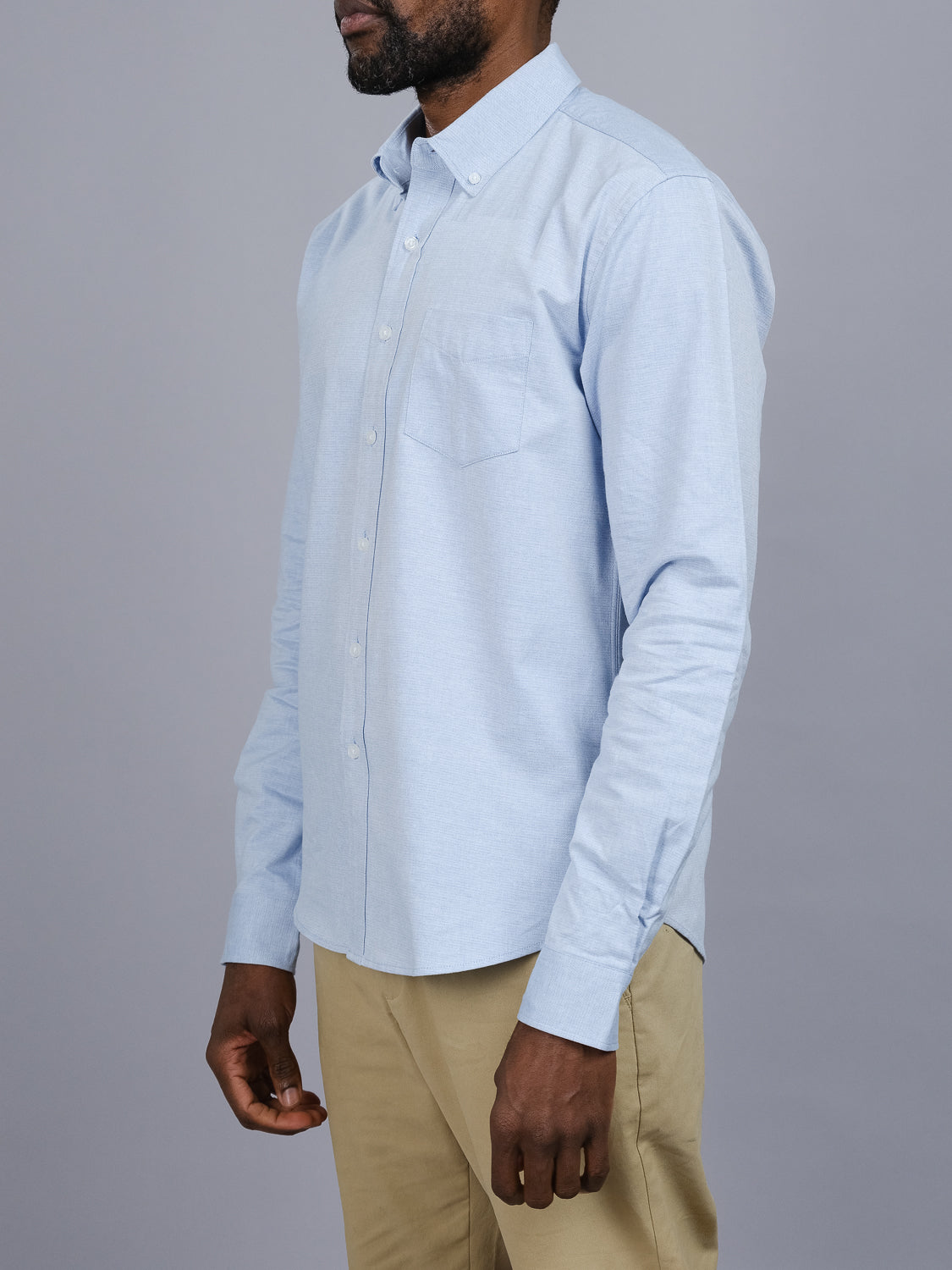 In front of a gray backdrop, someone stands slightly sideways wearing ID Menswears Akira Cotton Button Down Shirt in light blue. The shirt features a button-down collar, chest pocket, long sleeves, and is paired with beige trousers.