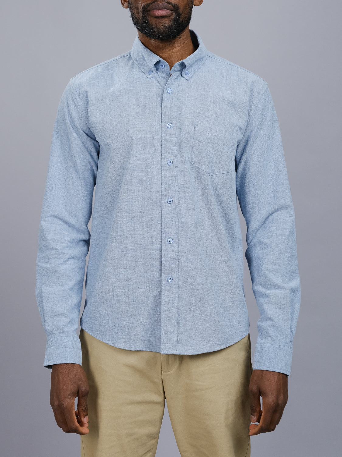 Wearing a light blue Akira Cotton Button Down Shirt by ID Menswear, featuring a regular fit and left chest pocket, a man pairs it stylishly with tan pants. He stands confidently against a plain gray backdrop.