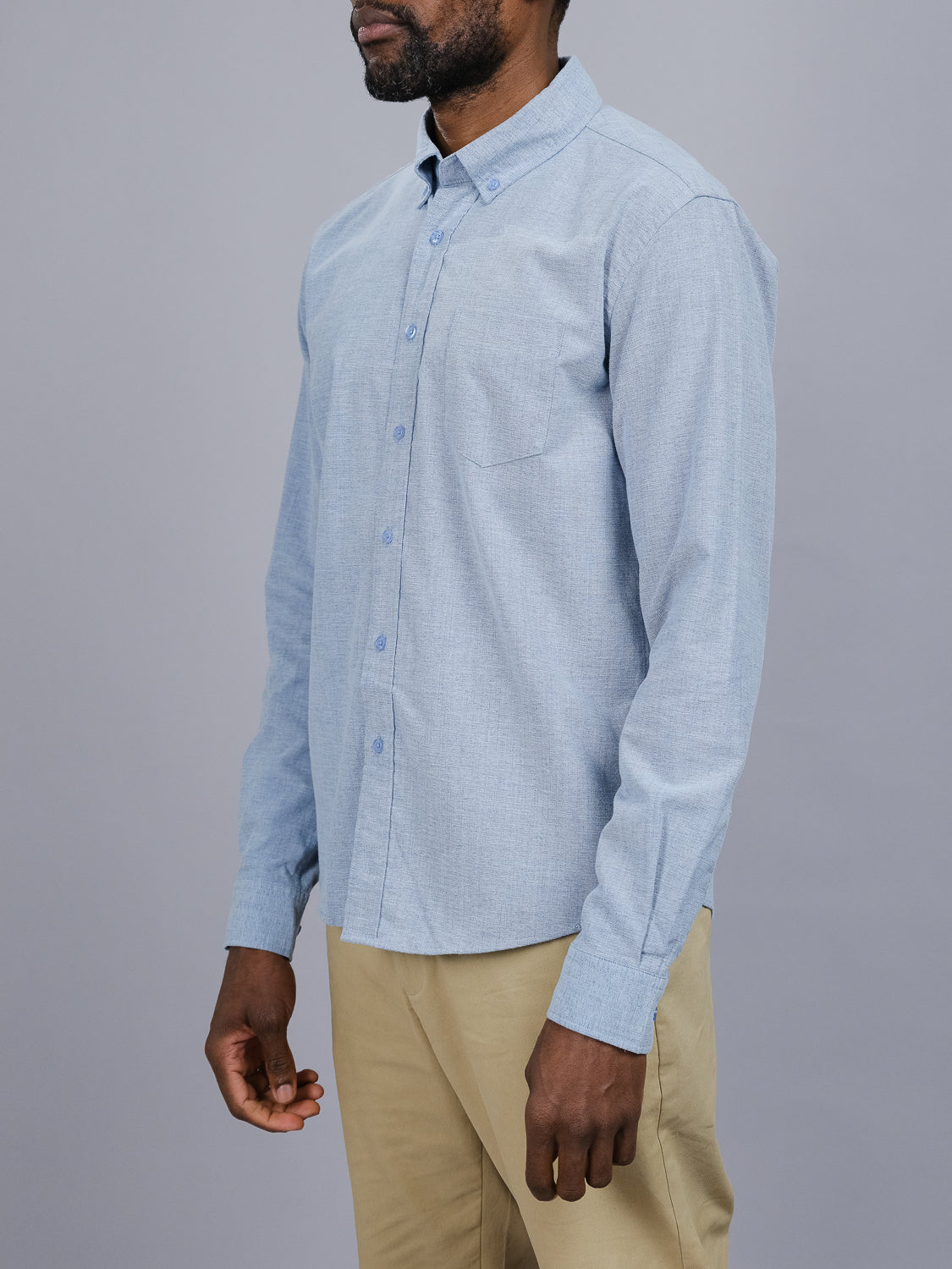 A bearded person stands against a gray background wearing an Akira Cotton Button Down Shirt in light blue and beige pants. This ID Menswear shirt features a left-side pocket and button-down collar, enhancing their relaxed demeanor as their hands rest by their sides.