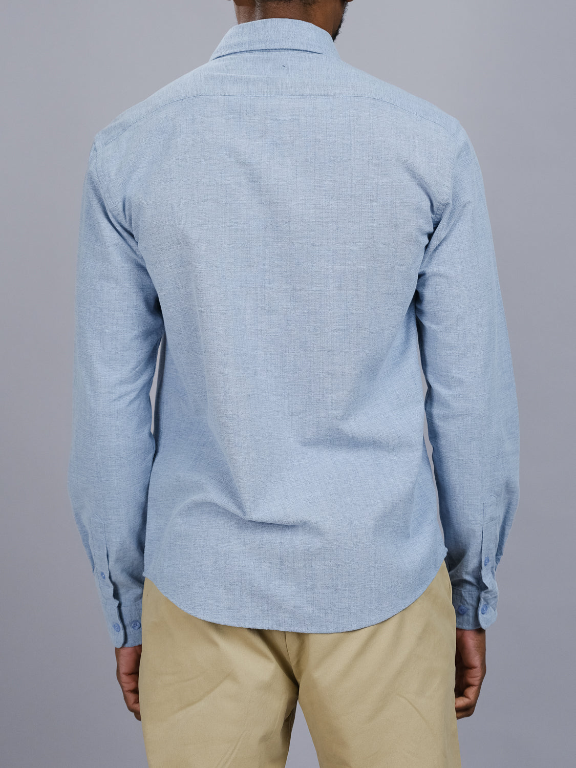 The rear view shows someone in a light blue Akira Cotton Button Down Shirt by ID Menswear, featuring buttoned cuffs and a button-down collar. Theyre paired with beige pants against a gray background, arms relaxed by their sides.