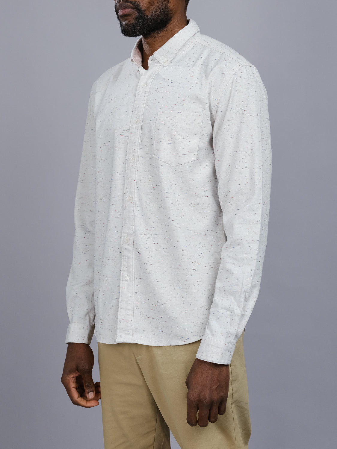 A person in an ID Menswear Allswell Speckled Button Down Shirt with tiny specks and khaki pants stands against a gray backdrop. The versatile design includes a left-side pocket, and they face slightly to the side.