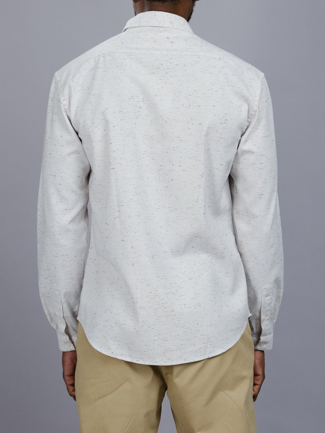 An individual in an Allswell Speckled Button Down Shirt by ID Menswear and beige pants stands facing away. The versatile cotton-linen design is highlighted against the plain gray backdrop, accentuating the light-colored ensembles subtle elegance.