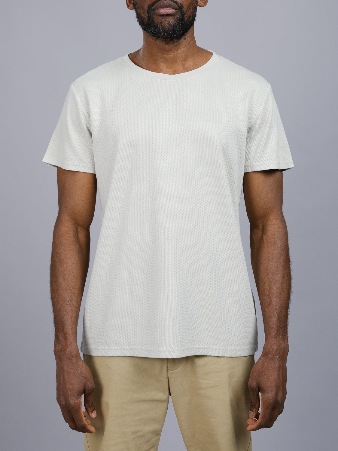 A man in an Urban Honeycomb Mesh Tee by ID Menswear, featuring a breathable bamboo-organic cotton blend and plain beige design, stands against a gray background with his khaki pants.