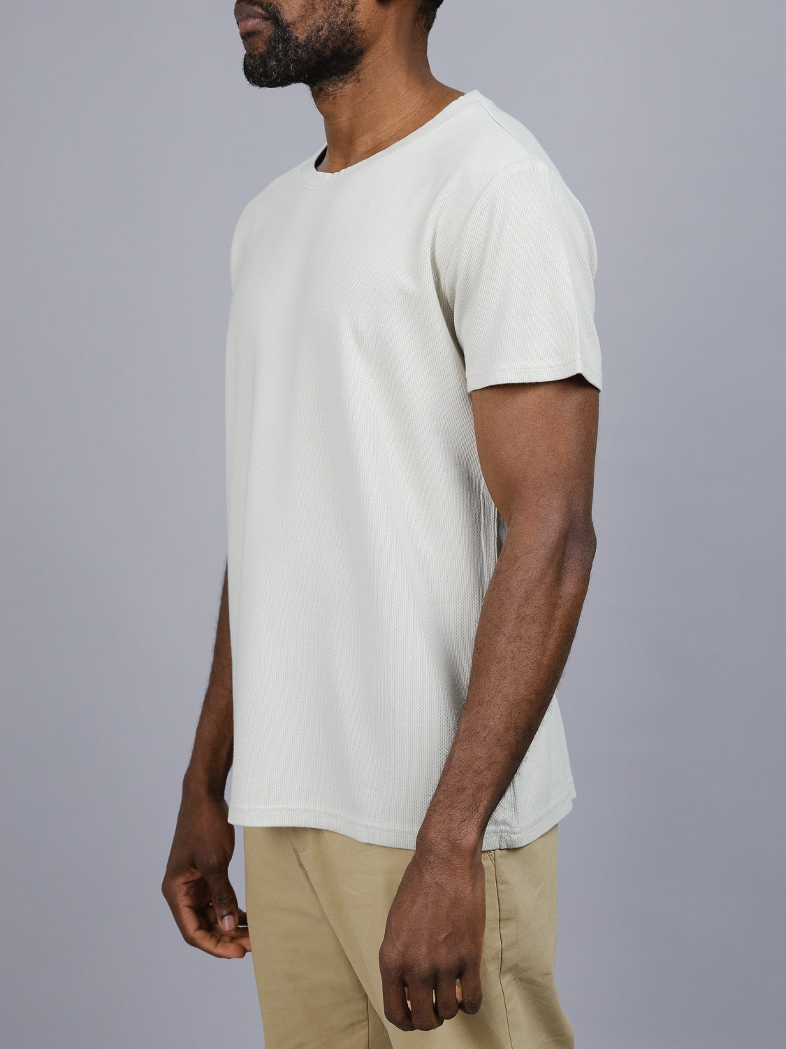 A man poses against a gray backdrop in the Urban Honeycomb Mesh Tee by ID Menswear, paired with beige pants made from a bamboo-organic cotton blend. He is turned slightly to reveal his side profile, with his arms relaxed at his sides.