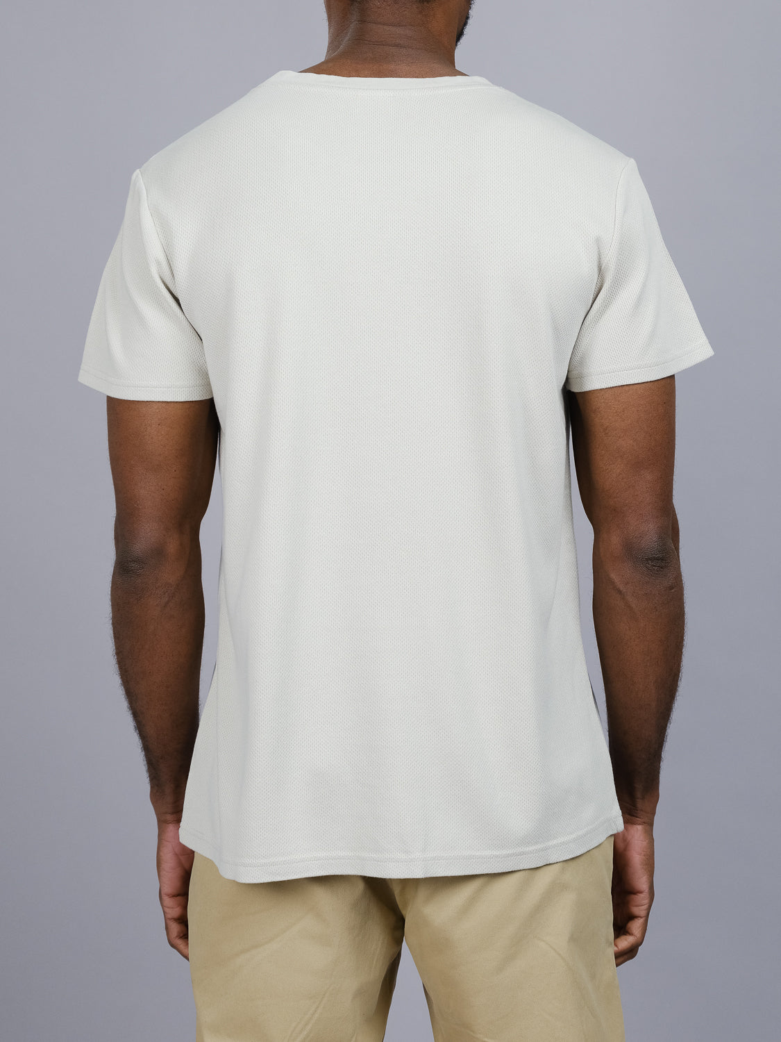 A person in a light gray Urban Honeycomb Mesh Tee by ID Menswear, featuring a relaxed fit and short sleeves, stands against a plain gray backdrop. They are also wearing beige pants.