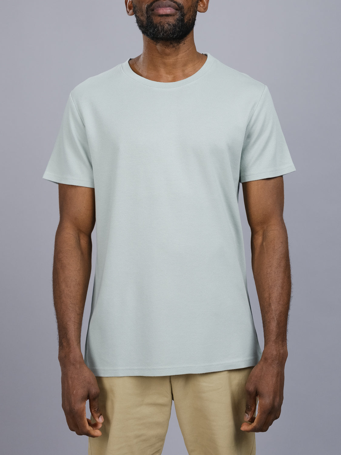An individual models the Urban Honeycomb Mesh Tee by ID Menswear, featuring its breathable, light gray honeycomb knitted mesh against a plain gray background. Only the short-sleeved, round-neck T-shirt and beige pants are visible in this cropped shot.