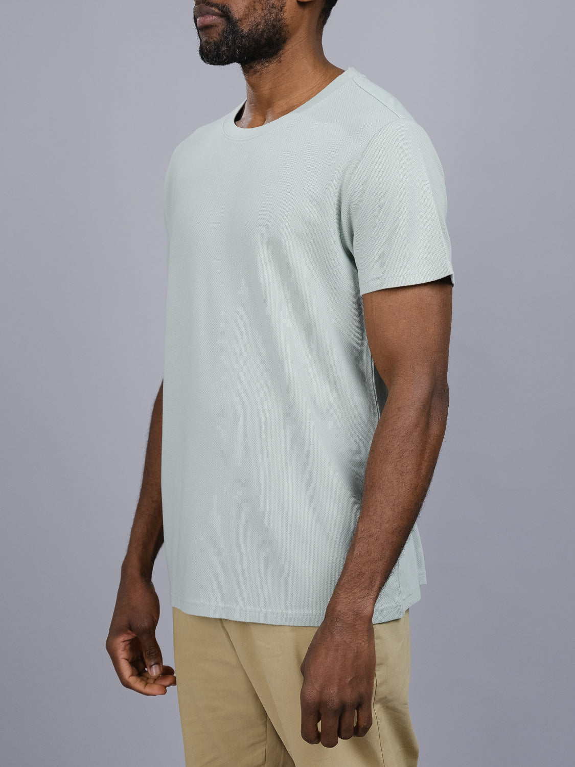 A person wears the Urban Honeycomb Mesh Tee in light gray by ID Menswear and beige pants against a plain gray background. Cropped from the neck down, the side profile shows their arms relaxed by their side.
