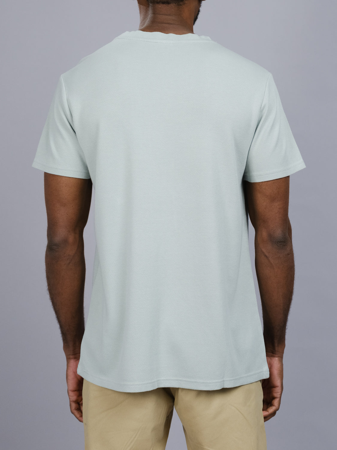 A person wears an Urban Honeycomb Mesh Tee in light gray by ID Menswear, made of a bamboo-organic cotton blend, paired with khaki pants. Captured from the back against a plain gray background, the image underscores the T-shirts simple design and breathable texture.