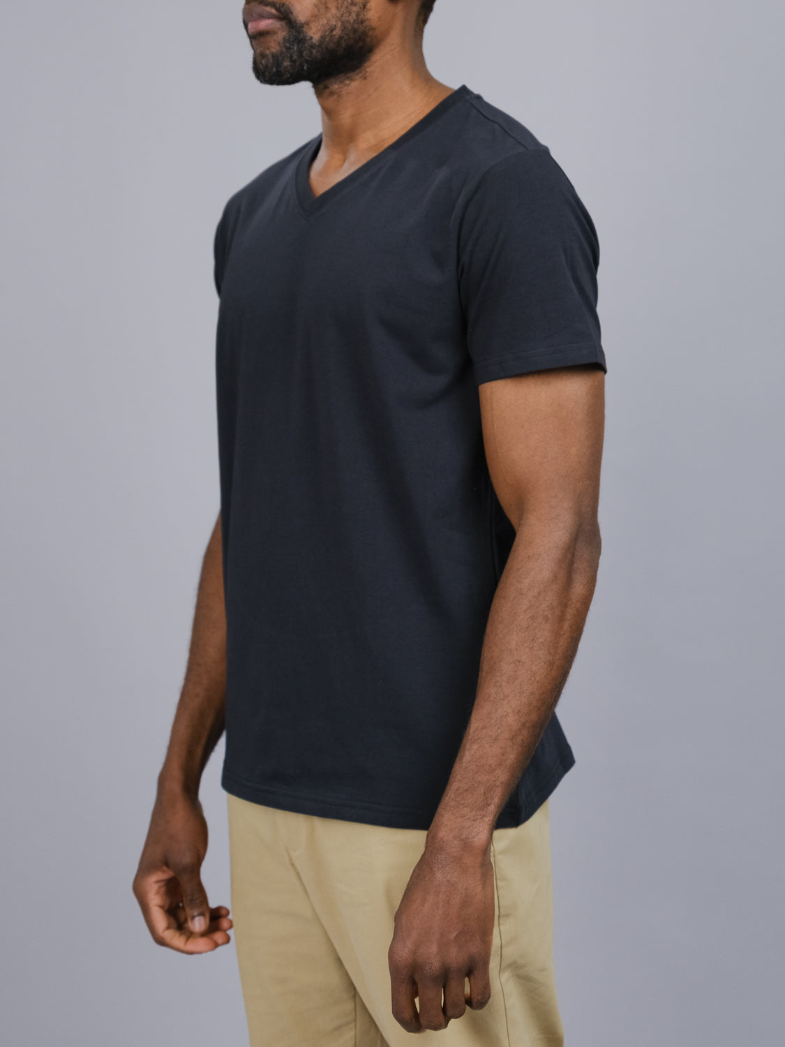 Against a gray background, a relaxed, bearded individual showcases effortless style wearing the black Classic V Neck Tee from ID Menswear paired with beige pants. This wardrobe staple, in a cotton-spandex blend, embodies comfort and elegance as they face slightly left.