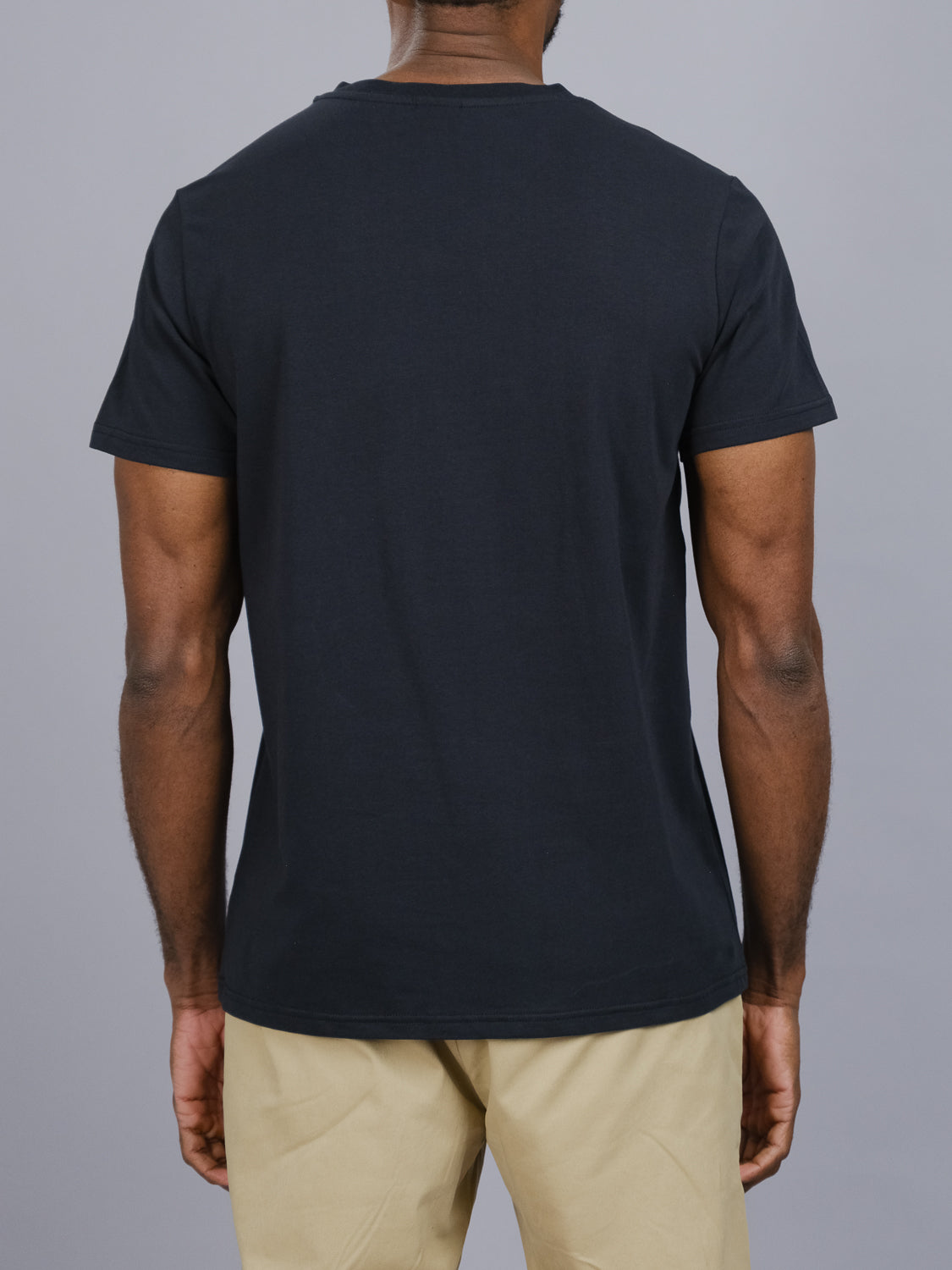 Back view of a person against a gray background, wearing an ID Menswear Classic V Neck Tee in black and beige pants. Crafted from a comfy cotton-spandex blend, this wardrobe essential complements the relaxed posture with arms at ease by their sides.