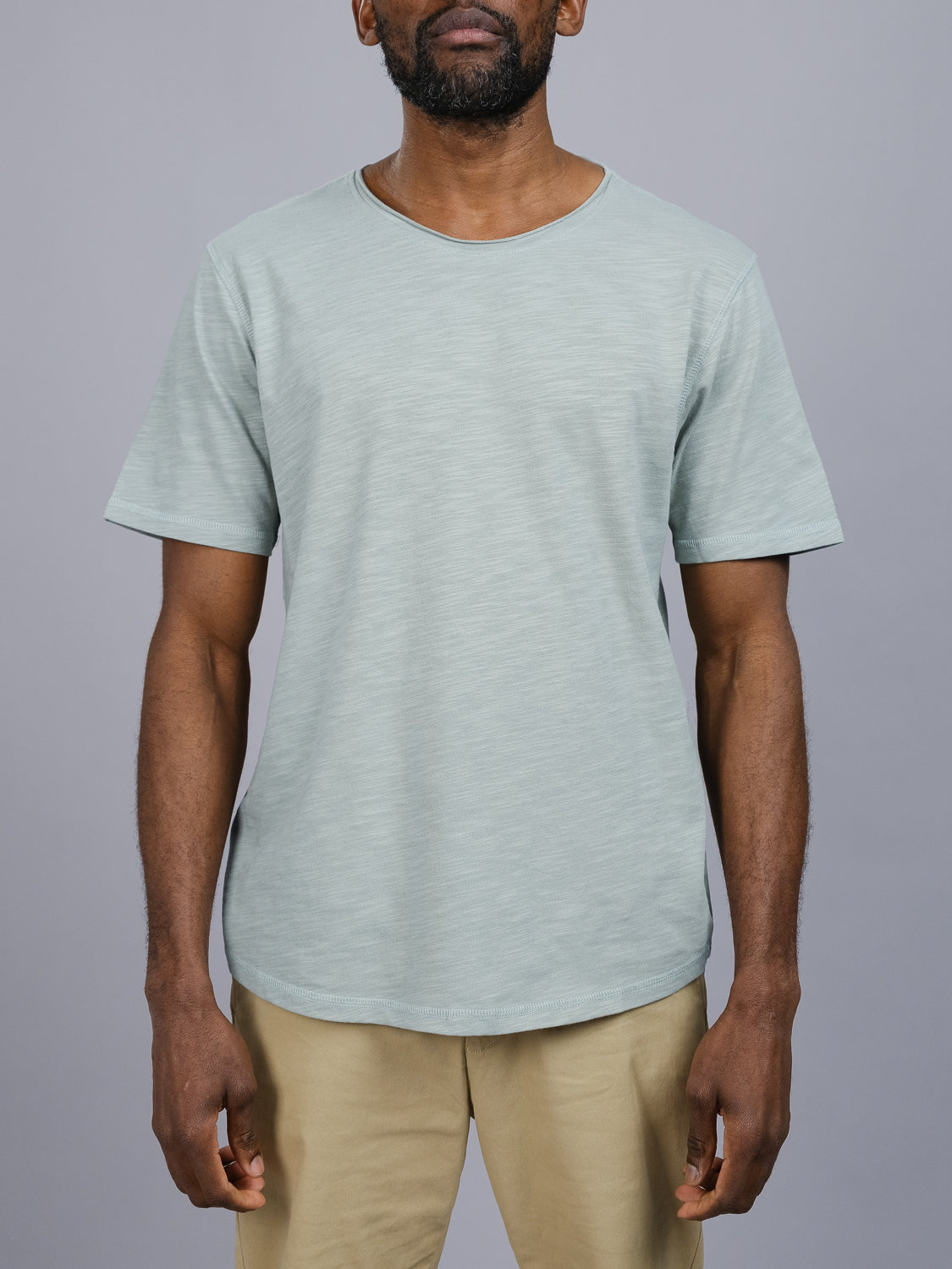 A man wears a Da Nang Rolled Neck Slub Tee by ID Menswear. The light green slub cotton tee features a rolled neck and subtle texture. He stands against a gray background in beige pants with his arms relaxed at his sides.