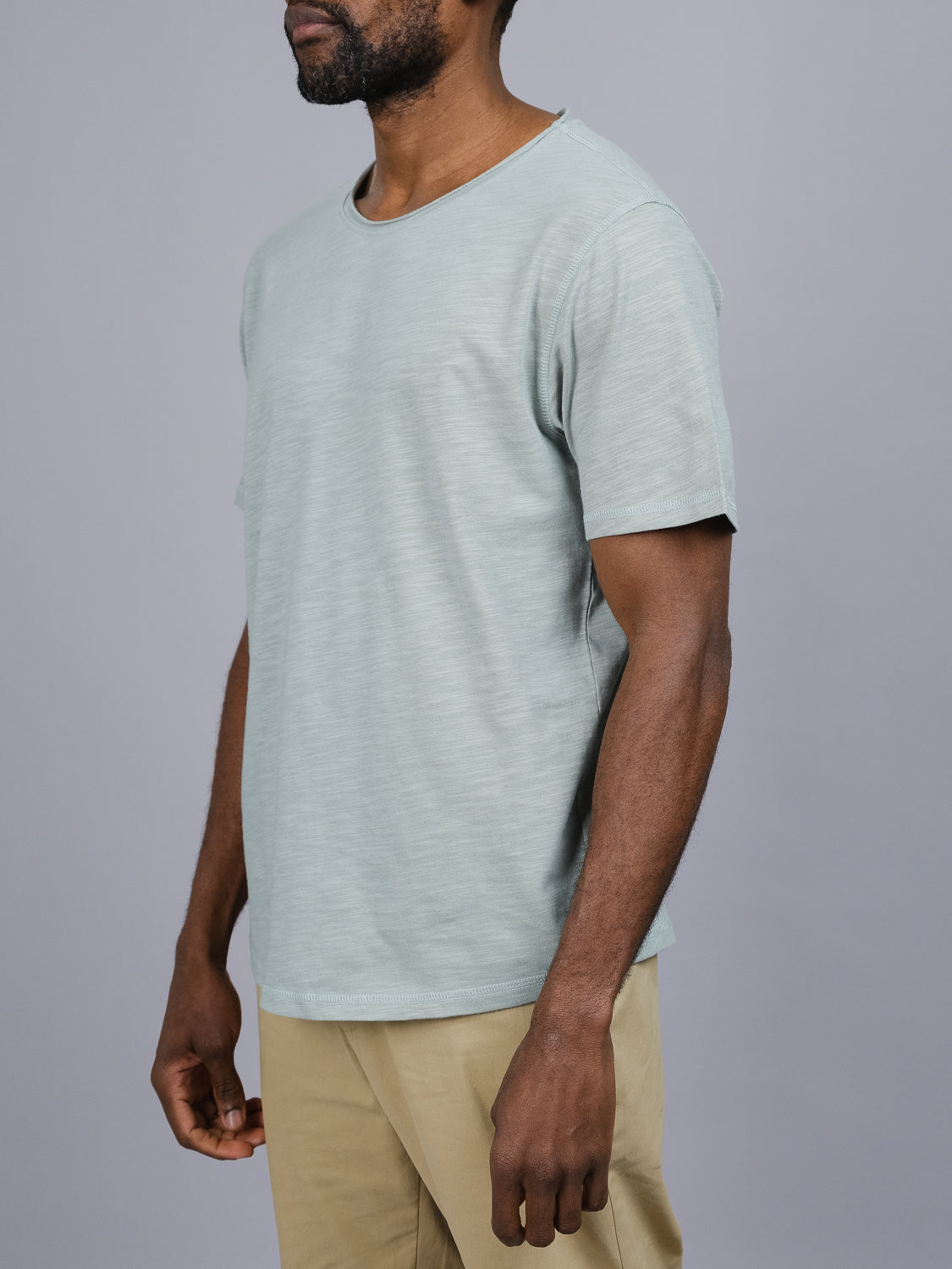 A man stands against a plain gray background in an ID Menswear Da Nang Rolled Neck Slub Tee in light green, paired with beige pants. Made of slub cotton, the tee has a slightly textured look. His face is not visible.