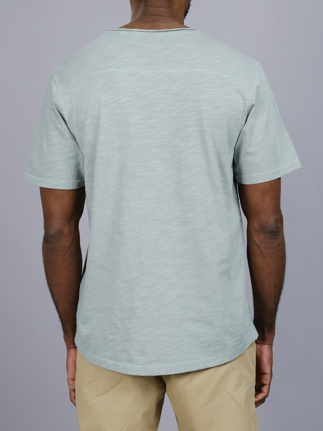 A person in an ID Menswear Da Nang Rolled Neck Slub Tee, a light blue cotton shirt with rolled neck short sleeves, and beige pants stands with their back to the camera against a plain gray background.