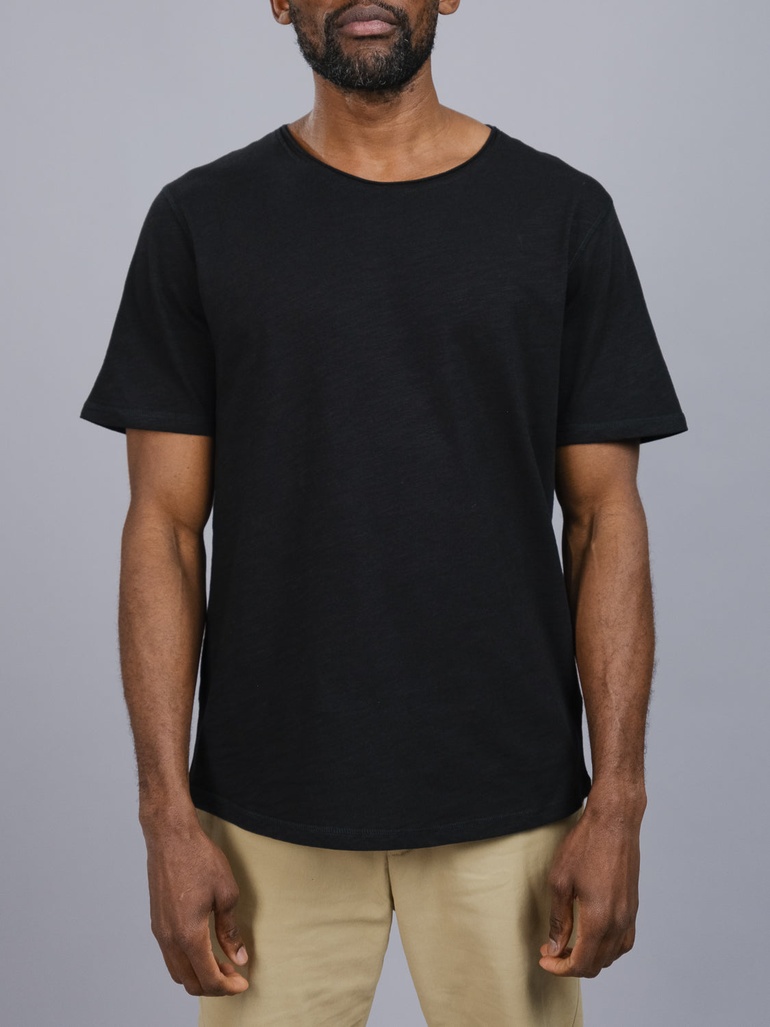 A man wears the Da Nang Rolled Neck Slub Tee by ID Menswear with beige pants against a gray backdrop. His arms rest casually at his sides while his face is out of frame, epitomizing effortless casual wear.
