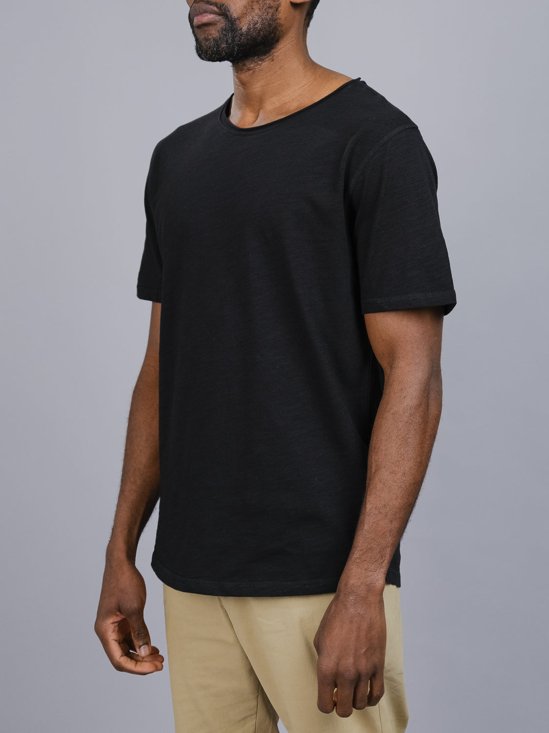 A man stands against a gray background, wearing the Da Nang Rolled Neck Slub Tee from ID Menswear and beige pants. His arms are relaxed at his sides as he gazes to the left.