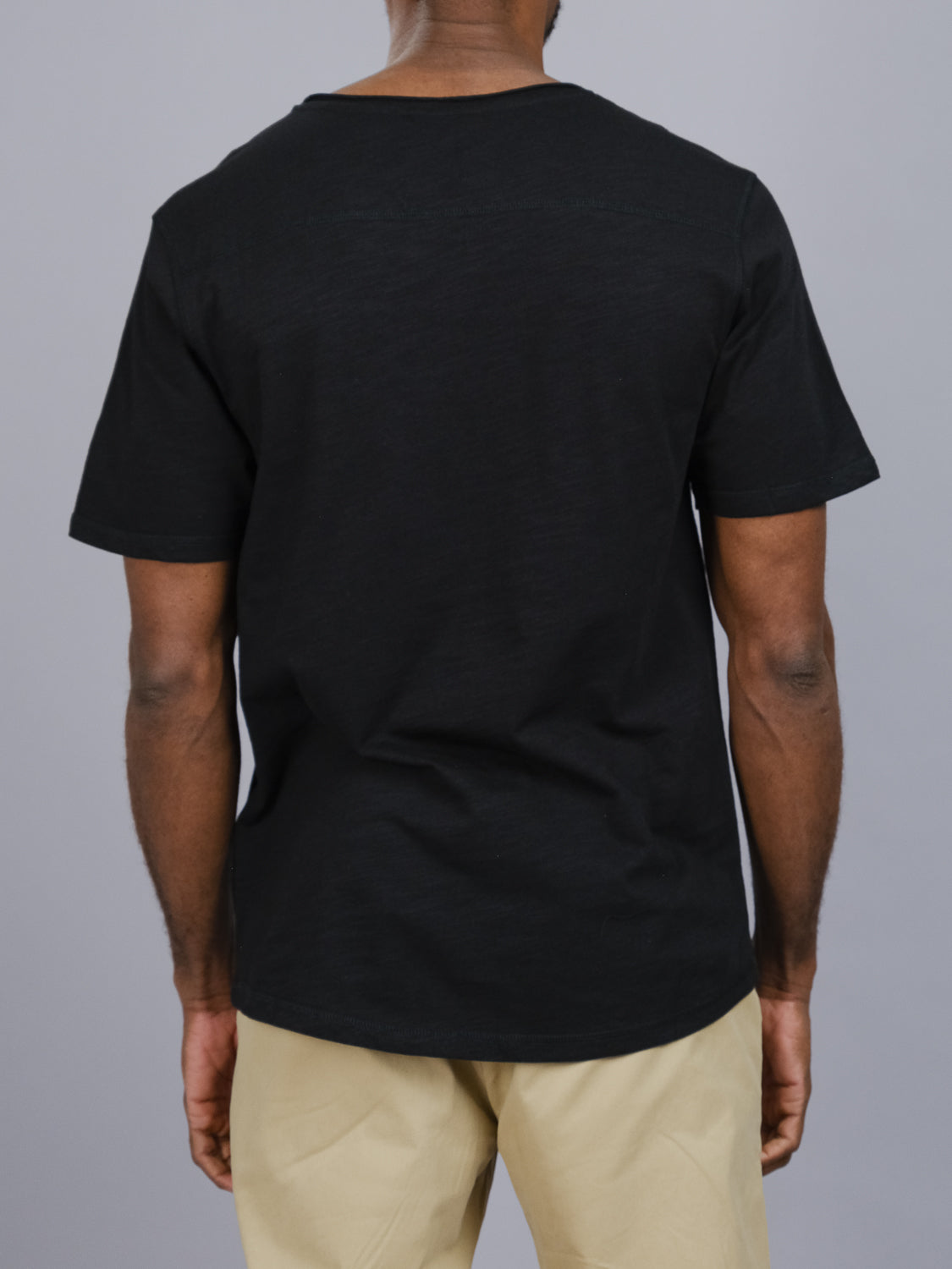 A person in casual attire is wearing an ID Menswear Da Nang Rolled Neck Slub Tee in plain black along with beige pants, facing away from the camera against a neutral gray backdrop.