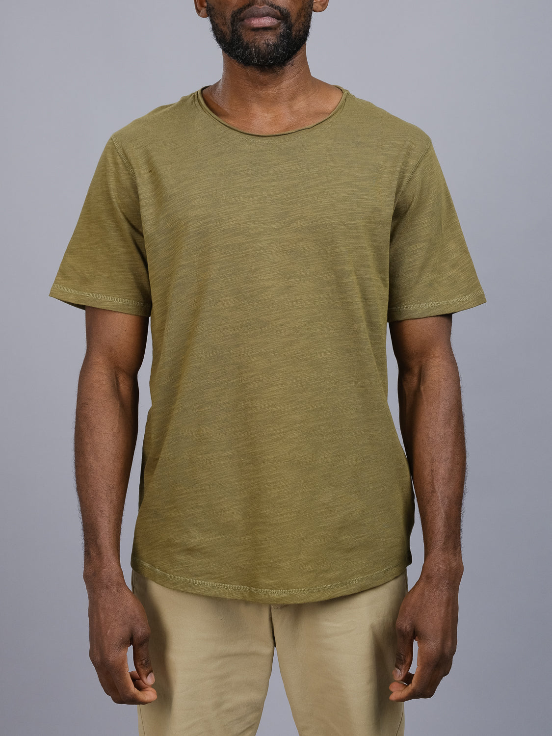 A person in an olive green Da Nang Rolled Neck Slub Tee by ID Menswear and beige pants stands against a plain gray backdrop. The T-shirt, with a round neckline and short sleeves, enhances the relaxed posture as their arms rest casually at their sides.