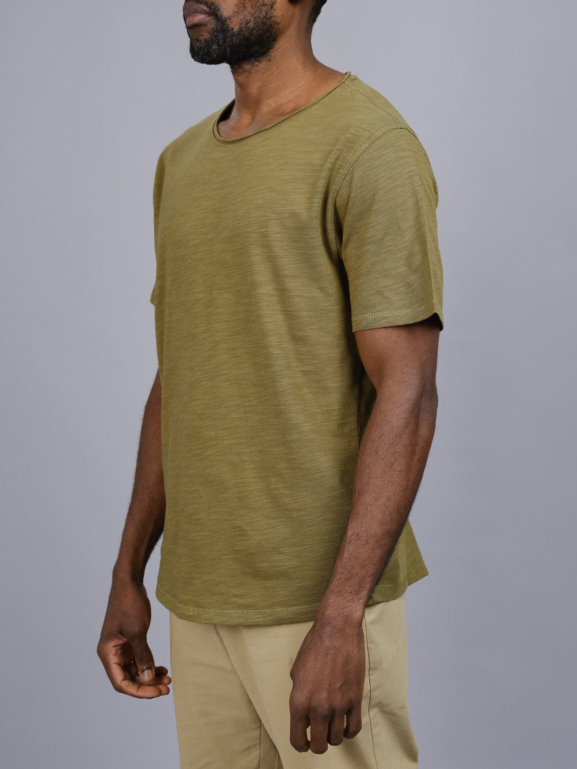 A person in an ID Menswear Da Nang Rolled Neck Slub Tee and beige pants stands against a gray background. The individual faces slightly to the side, arms relaxed at their sides, with the image focusing on the upper body.