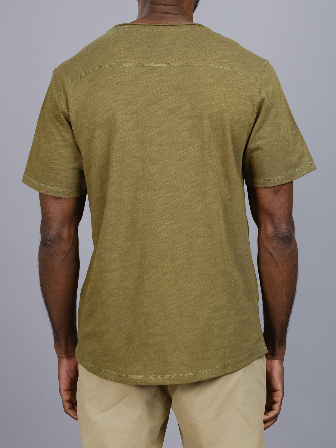 A person wearing the Da Nang Rolled Neck Slub Tee from ID Menswear in green and beige pants stands facing away from the camera against a plain gray background, showcasing the tees textured fabric for added depth.
