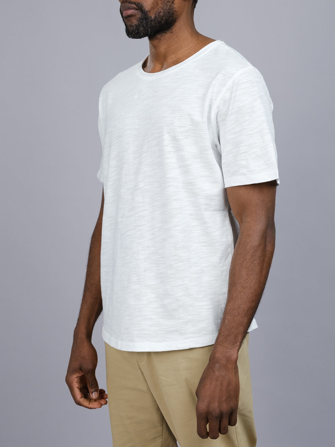 A person is seen from the side, wearing an ID Menswear Da Nang Rolled Neck Slub Tee and beige pants against a plain gray background, with their face out of view to emphasize the effortless style of the tees rolled neck design.