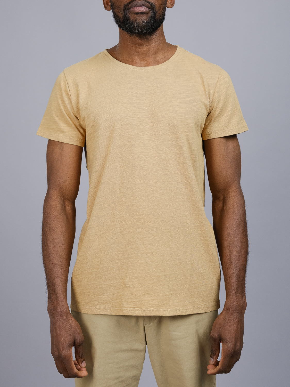 A person stands facing the camera in a Mini Flip Slub Yarn Tee and shorts by ID Menswear, set against a plain gray background. Their arms are relaxed, creating an effortless casual look. The setting is simple and neutral, ideal for everyday wear.