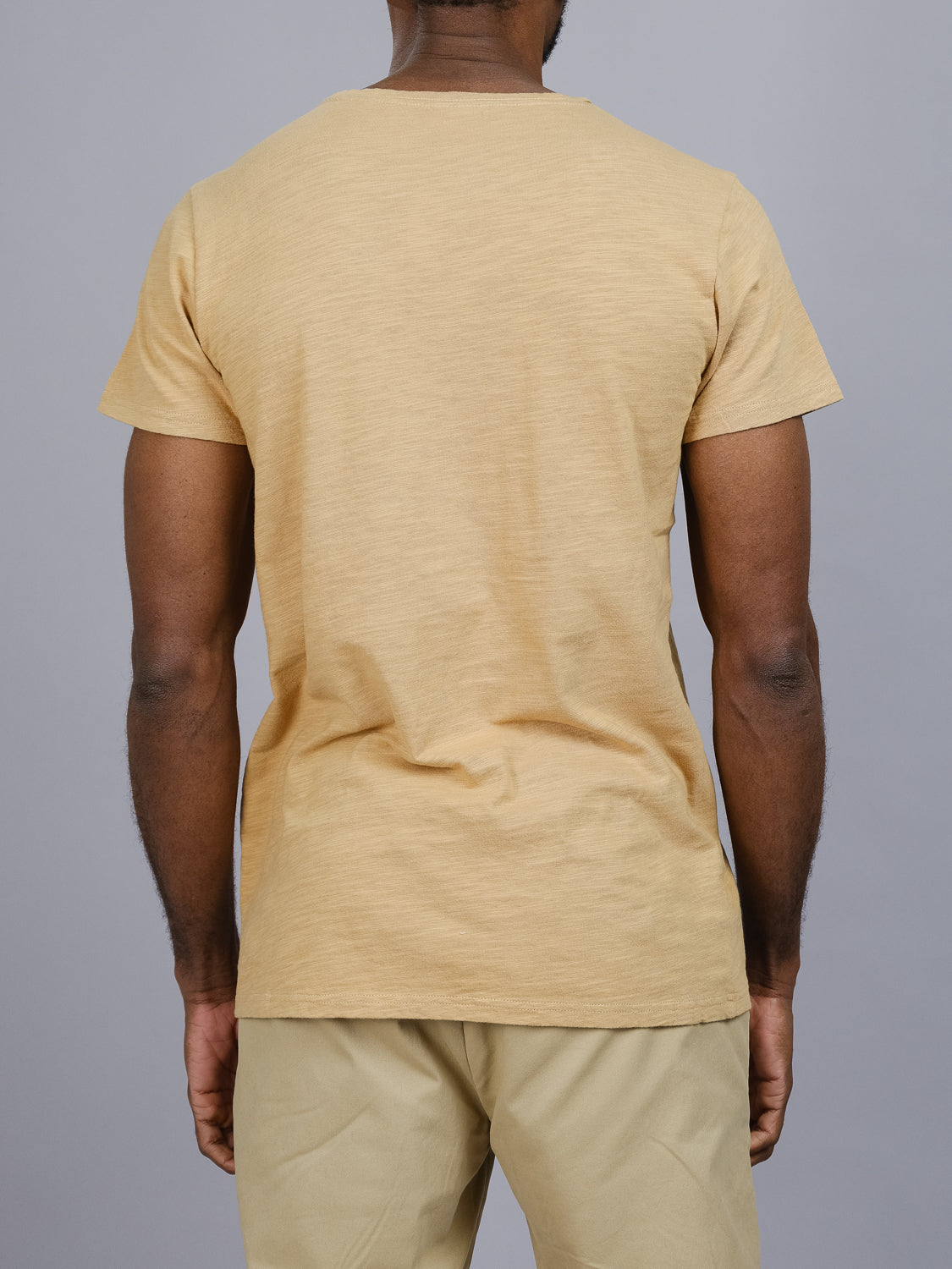 A person wearing the Mini Flip Slub Yarn Tee by ID Menswear in beige and light-colored pants stands against a plain gray backdrop, embodying an effortlessly casual look.