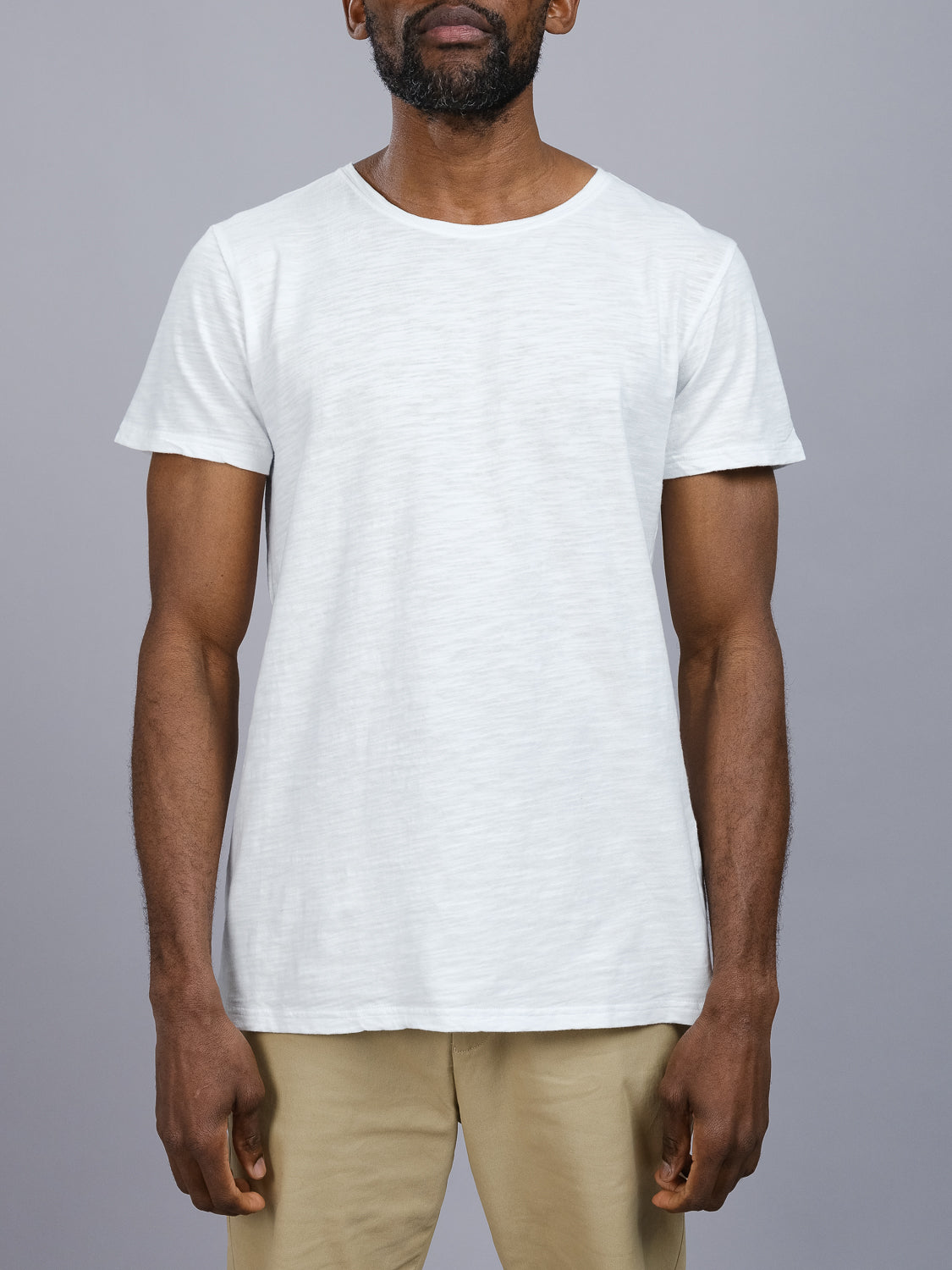 A person in an ID Menswear Mini Flip Slub Yarn Tee and beige pants stands against a gray background, embodying everyday wear with casual style. Their arms rest naturally at their sides, showcasing effortless fashion.