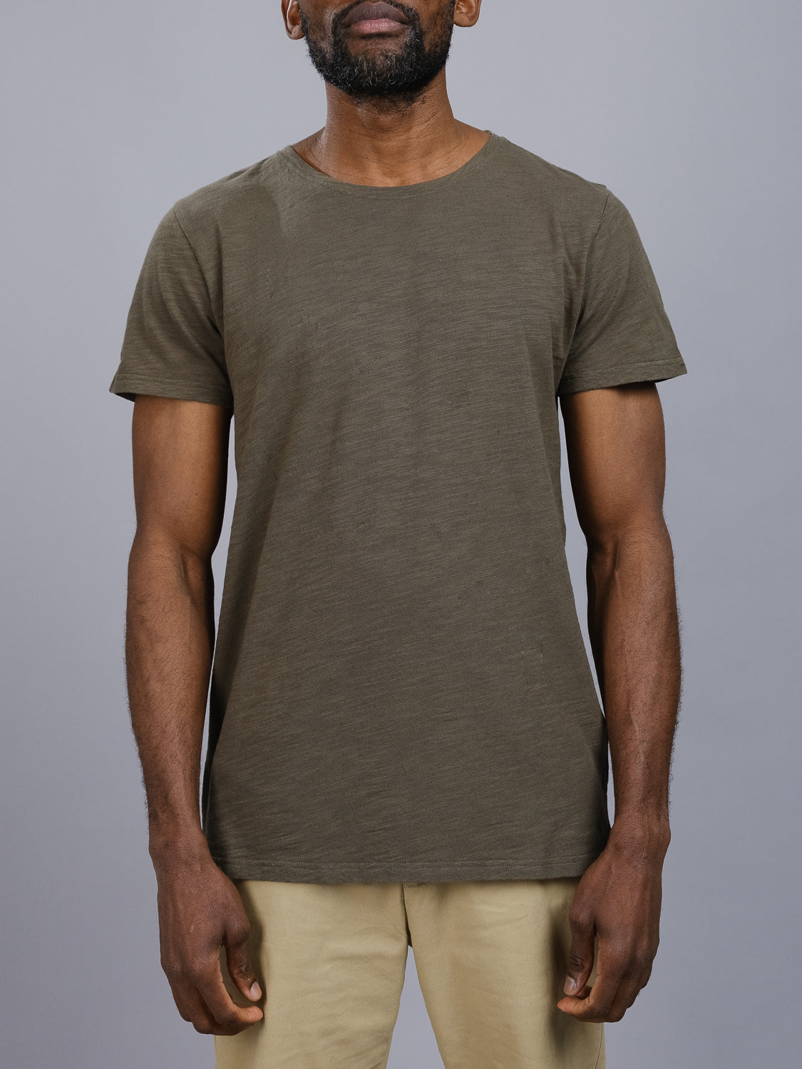 A man in a relaxed, casual look sports an olive Mini Flip Slub Yarn Tee from ID Menswear with beige pants against a gray backdrop, his calm demeanor reflected in his arms resting by his sides, perfectly epitomizing understated everyday style.