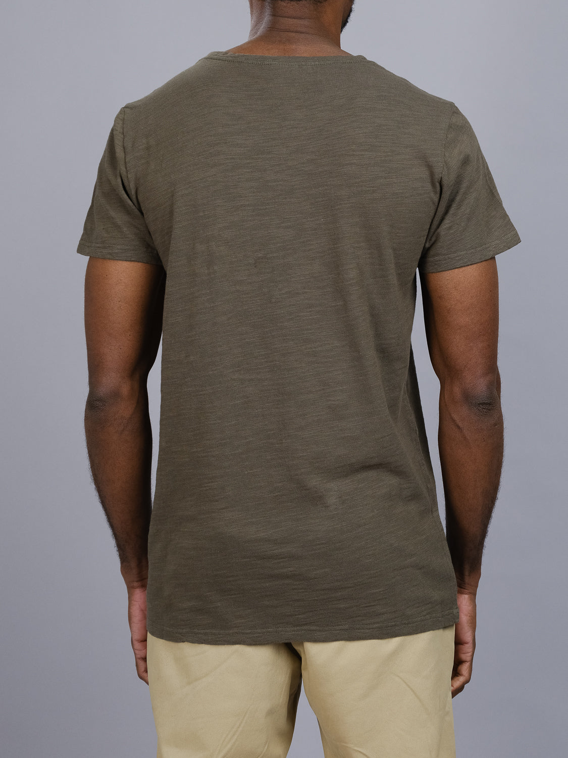 A person wears an ID Menswear Mini Flip Slub Yarn Tee in plain olive green paired with beige pants, facing away from the camera against a gray background.