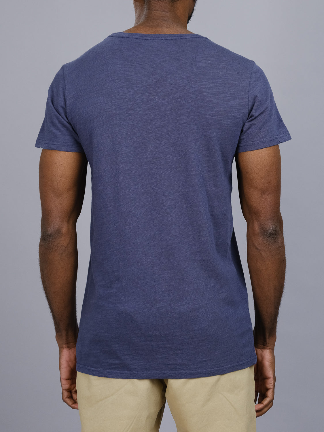 Dressed casually in an ID Menswear Mini Flip Slub Yarn Tee, a person wearing a solid dark blue slub cotton T-shirt and beige pants stands with their back to the camera against a plain gray background.