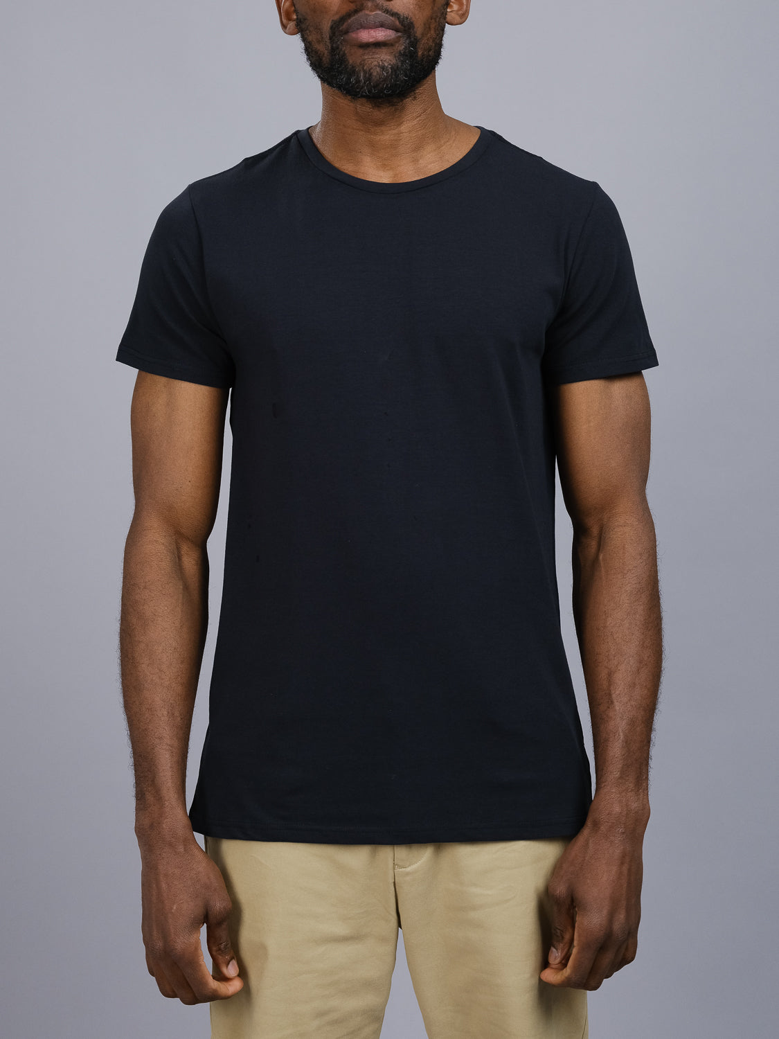 A person stands against a gray background, wearing a Form Crew Neck Tee by ID Menswear. The outfit includes a form-fitting black T-shirt and beige pants, both made from comfortable fabric. The persons face remains unseen.